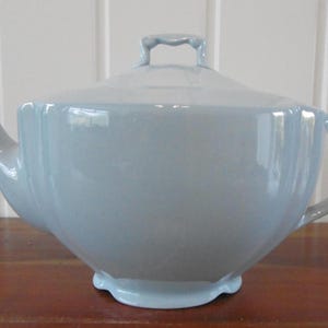 May include: A pale blue ceramic teapot with a lid and handle. The teapot has a curved spout and a rounded body. The handle is arched, and the lid has a small handle. The teapot is approximately 25 cm wide and 15 cm tall.