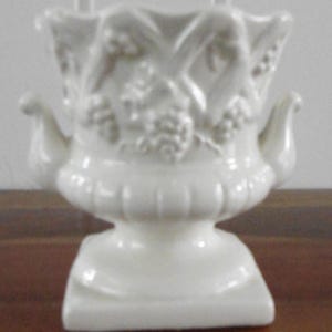 Puede incluir: Una urna de cerámica blanca con base cuadrada y cuerpo bulboso. La urna presenta detalles en relieve de uvas y hojas, con asas curvas a cada lado. La parte superior tiene un borde decorativo festoneado. Una pieza decorativa clásica.