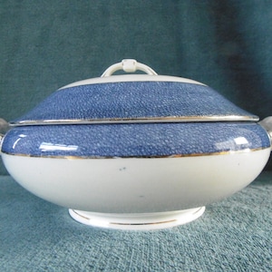 May include: A white porcelain serving dish with a blue and gold rim. The dish has a lid with a knob on top. The dish is oval shaped and has two handles on the sides.