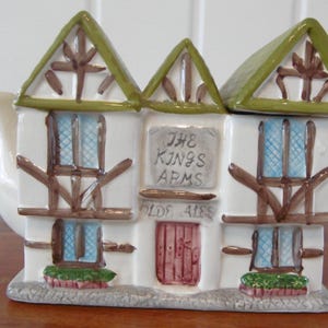 May include: White ceramic teapot shaped like a building with green roofs and brown trim. The sign reads "The Kings Arms Olde Ales".