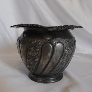 May include: A dark metallic bowl with a wide, scalloped rim featuring intricate floral and scrollwork engravings. The body of the bowl has vertical fluted panels alternating with detailed embossed patterns, resting on a circular pedestal base.