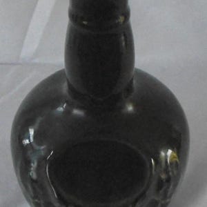 May include: A dark green glass bottle with a round body and a narrow neck. The bottle has a textured surface and a circular opening at the top.