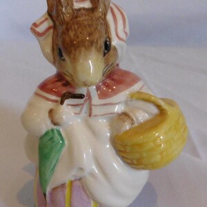 Peut inclure: Figurine en céramique représentant un lapin vêtu d'un bonnet et d'une robe à rayures blanches et rouges. Le lapin tient un panier jaune et un parapluie vert. La base est verte avec une jupe rayée rose et jaune.