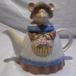 Könnte beinhalten: Eine skurrile Teekanne mit einem Mäusefigurendeckel, die einen Hut und ein Kleid trägt. Die Teekanne ist cremefarben mit blauer Bordüre und einem Korb mit rosa Rosen. Die Maus ist braun und beige.