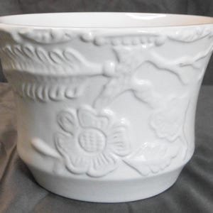 May include: A white ceramic plant pot with embossed floral and leaf patterns. The pot has a gently flared shape and a detailed, textured surface. The embossed designs include flowers, leaves, and other decorative elements, enhancing the visual appeal of the item.
