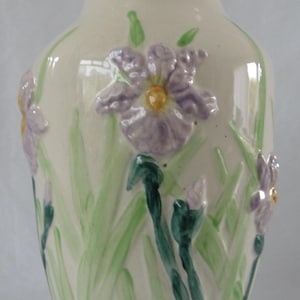 May include: Cream ceramic vase featuring embossed purple iris flowers and green leaves. The vase has a wide base, a cylindrical body, and a flared rim. The iris flowers have yellow centres.