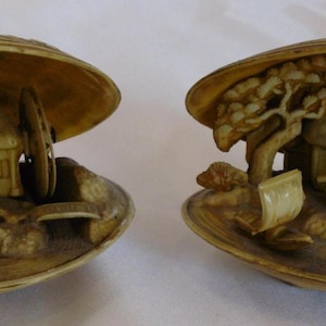 Könnte beinhalten: Zwei dekorative Muschelschalen, jede mit einer Miniaturszene. Die Szenen zeigen ein kleines Haus, einen Baum und Figuren, alles in einem hellbeigen Farbton geschnitzt. Die Muscheln sind geöffnet und zeigen die detaillierten Innenschnitzereien.
