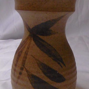 May include: A tan ceramic vase with a wide top and a narrow neck. The vase features dark brown leaf-like designs. The vase has a textured surface and a rounded base. The vase is likely handmade and suitable for home decor.