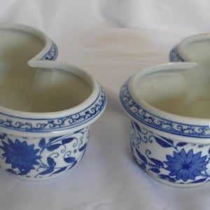 May include: Two ceramic clover-shaped bowls featuring a white base with blue floral patterns and scrolling vines. Each bowl has a thick, rounded rim decorated with a blue and white geometric border, and a smooth, off-white interior finish.