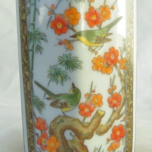 Può includere: Un vaso rettangolare in ceramica con un design di ispirazione asiatica. Il vaso presenta uno sfondo bianco con una dettagliata illustrazione di uccelli verdi, fiori arancioni e bambù. I bordi sono rifiniti con un bordo dorato.