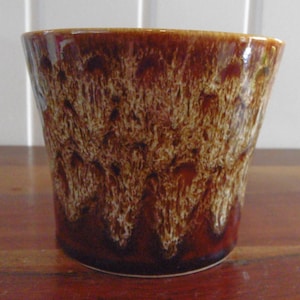 May include: A brown ceramic planter with a textured, speckled design. The planter has a wide opening and a slightly tapered shape. The glaze has a glossy finish, with a darker brown base that fades into lighter brown and cream tones.