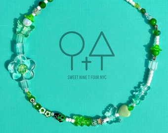Op de afbeelding: Een groene en witte kralenketting met verschillende bedels, waaronder bloemen, vlinders, sterren en een hart. De ketting heeft een zilveren sluiting en bevat de tekst "O+T SWEET NINE T FOUR NYC".