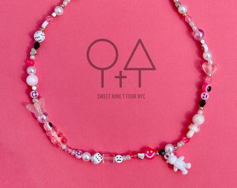 Op de afbeelding: Een roze en witte kralenketting met verschillende bedels, waaronder harten, dobbelstenen, smiley-gezichten en een vlinder. De ketting heeft een zilveren sluiting en bevat de tekst "O+T SWEET NINE T FOUR NYC".