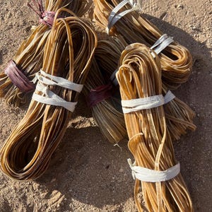 Puede incluir: Cinco paquetes de tiras de cuero marrón atadas con cuerda blanca y morada. Las tiras de cuero están enrolladas y apiladas unas sobre otras.