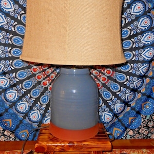May include: A blue ceramic table lamp with a burlap shade. The lamp has a rounded base with a terracotta-coloured bottom. It sits on a small, wooden, three-legged table. The background features a patterned fabric.