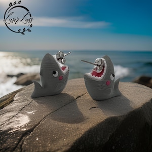 May include: A pair of gray shark earrings with open mouths and pink cheeks. The earrings are made of polymer clay and have silver posts.