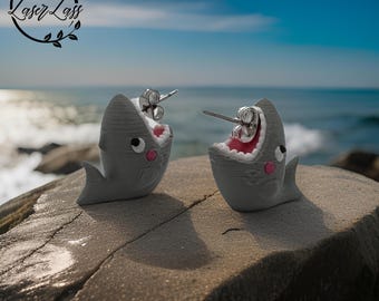May include: A pair of gray shark earrings with open mouths and pink cheeks. The earrings are made of polymer clay and have silver posts.