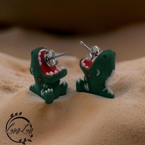 Pode incluir: Um par de brincos de botão em forma de dinossauro verde com bocas vermelhas e dentes brancos. Os brincos têm pinos de prata. Um acessório divertido e divertido.