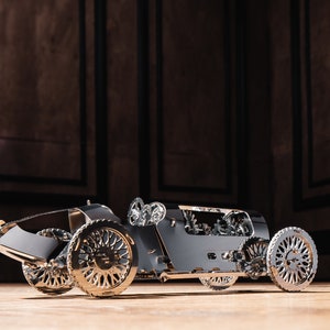 Könnte beinhalten: Ein silbernes Modellauto mit filigranen Details, darunter sichtbare Zahnräder und Räder. Das Auto steht auf einer Holzoberfläche mit einem dunklen Holzhintergrund.