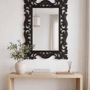 May include: A rectangular wall mirror featuring a thick, matte black frame with intricate, symmetrical scrollwork and carved leaf-like patterns along all four sides. The mirror glass is clear and reflects the surrounding room interior.