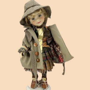 May include: A doll dressed in a tan trench coat, plaid hat, and a patterned skirt. The coat has large brown buttons and a plaid accent. The doll wears brown shoes and a yellow tie. The doll has blonde hair and is standing on a white base.
