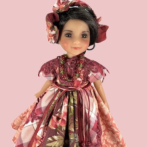 May include: A doll wearing a red and white plaid dress with floral accents. The dress has a lace collar and a red ribbon tied around the waist. The doll has brown hair and brown eyes and is wearing a red floral headband.