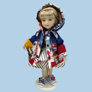 May include: A doll wearing a patchwork jacket with a hood, a striped dress, and matching sneakers. The jacket is primarily navy blue with red, yellow, and patterned fabric accents. The doll has blonde hair and is standing on a white stand.