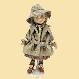 May include: A doll dressed in a tan trench coat with a matching hat, brown shoes, and a plaid skirt. The coat has a plaid collar and belt. The doll is standing on a white stand.