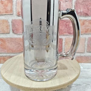 May include: A clear glass beer mug with a silver handle and vertical stripes. The mug features etched text that reads "Hiram's Ri'" and "Pigeon Forge". The mug is displayed on a small wooden stand against a brick background.