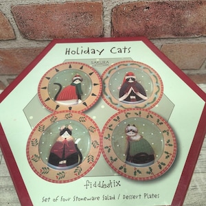 May include: A set of four Sakura Holiday Cats stoneware salad or dessert plates in a hexagonal box. Each plate features a cat wearing winter clothing, including scarves and hats, against a light green background with white dots and holly borders.