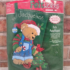 May include: Felt applique kit for a Christmas stocking featuring a bear wearing a Santa hat and overalls, holding a candy cane and a bird. The stocking is approximately 18 inches (46 cm) long and is labeled "Jacqueline."