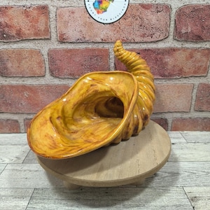 May include: A decorative, yellow and brown ceramic cornucopia, resting on a small, round wooden stand with three legs. The cornucopia has a curved, horn-like shape and a textured surface. The background features a brick wall.