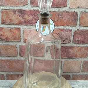 May include: A clear glass decanter with a decorative stopper. The decanter has a square shape with beveled edges and a cork stopper topped with a cut-glass design. The decanter is displayed on a wooden surface against a brick wall.