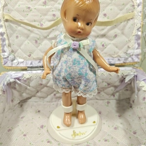 May include: A vintage doll with a light brown complexion, wearing a blue floral romper with white lace trim. The doll has a bow with a purple rose accent. It stands on a white base inside a floral-lined case.