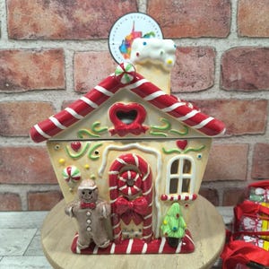 May include: A ceramic gingerbread house cookie jar with red and white striped roof and accents. Features a gingerbread man, candy cane door, and Christmas tree. Set on a wooden stand.