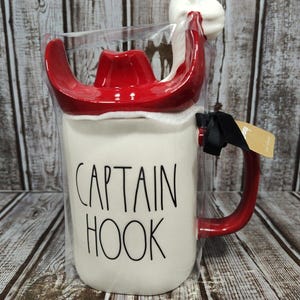 May include: White ceramic mug with a red handle and a red Santa hat lid. The mug has the text "CAPTAIN HOOK" printed in black.