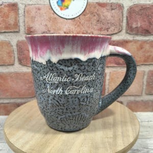 May include: A speckled gray ceramic mug with a dark pink rim and the words "Atlantic Beach North Carolina" in white script. The mug has a curved handle and sits on a wooden stand.