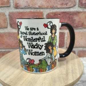 May include: White ceramic mug with a black handle and rim. The mug features an illustration of women and the text "We are a Sacred Sisterhood of Wonderful Wacky Women". The mug is on a wooden surface.