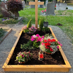 May include: A wooden cross is positioned above a rectangular flower bed filled with vibrant flowers and greenery. A small glass lantern with a lit red candle is placed near the flowers. The setting is a cemetery.