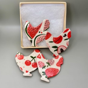 May include: Four ceramic chicken-shaped ornaments with fruit designs. The chickens are white with red, pink, and green fruit patterns, including watermelon, cherries, and strawberries. One chicken is inside a small, open cardboard box.
