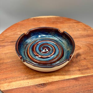 以下が含まれることがあります： 青と茶色のスパイラルデザインの手作りセラミック灰皿。 灰皿は白いベースと濃い茶色の縁があります。 丸くて装飾的な作品です。