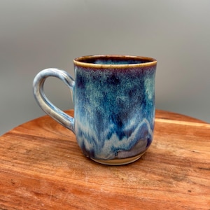 Könnte beinhalten: Eine handgefertigte Keramiktasse mit blau-weißer Glasur. Die Tasse hat einen abgerundeten Körper, einen gebogenen Henkel und einen braunen Rand. Die Tasse steht auf einer Holzoberfläche.