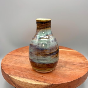 May include: A small, handcrafted ceramic vase with a rounded body and a narrow neck. The vase features a blend of colours, including shades of brown, blue, and green. It is displayed on a round, wooden stand with black metal legs.