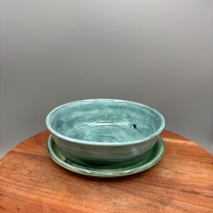 Puede incluir: Un juego de cuenco y platillo de cerámica hechos a mano. El cuenco es de color verde azulado apagado con una superficie texturizada y un pequeño orificio de drenaje. El platillo es verde a juego. El juego se exhibe sobre una superficie de madera.