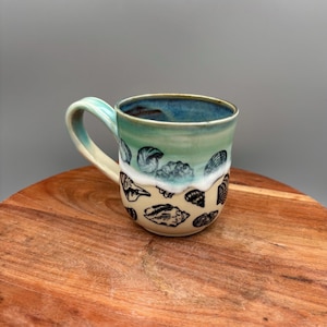 Puede incluir: Una taza de cerámica con un diseño de temática marina. La taza tiene un interior azul claro y un asa a juego. El exterior está decorado con un degradado de colores, que va del azul claro al blanco y luego a una base beige con ilustraciones de conchas negras.