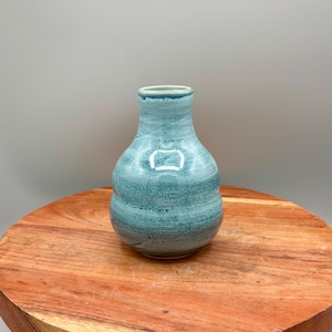 May include: A small, turquoise ceramic vase with a textured, banded design. The vase has a rounded body and a narrow neck. The vase is displayed on a round, wooden stand.