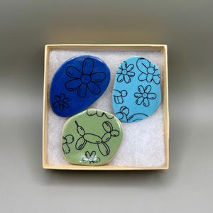 May include: A set of three decorative ceramic stones in a small box. The stones are oval-shaped and feature hand-drawn designs. One is blue with a flower, another is light blue with flowers, and the third is green with a balloon dog.