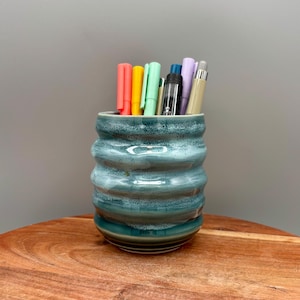 May include: A teal ceramic pen holder with a textured, ridged design. The holder contains a variety of colorful pens and markers. The pen holder sits on a wooden surface against a gray background. The ceramic has a glossy finish.
