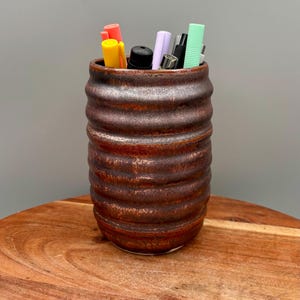 Puede incluir: Un portalápices de cerámica marrón con un diseño texturizado y acanalado. El portalápices está lleno de varios bolígrafos y rotuladores de colores. El portalápices está sobre una superficie de madera.