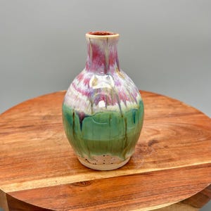 Könnte beinhalten: Eine kleine, handgefertigte Keramikvase mit rundem Körper und schmalem Hals. Die Vase hat eine lebendige Farbpalette mit einer Mischung aus Rosa, Weiß, Grün und Lila. Die Vase wird auf einer Holzoberfläche präsentiert.